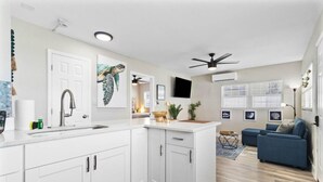Une cuisine moderne avec des meubles blancs, un évier et un ventilateur de plafond. On y trouve également un coin repas avec un canapé bleu et une petite table.