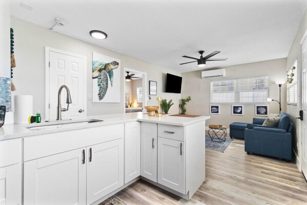 A modern kitchen with white cabinets, a sink, and a ceiling fan. There is a dining area with a blue sofa and a small table.