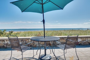 Une table ronde en métal, accompagnée de deux chaises et d’un grand parasol, est placée devant un mur de pierre, avec vue sur une étendue d’eau.
