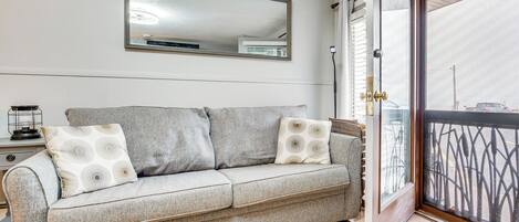 Een woonkamer met een grijze bank, twee kussens met een patroon, een grote spiegel en een schuifdeur naar buiten.