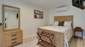 A bedroom with a bed, a wooden dresser, a mirror, a wall-mounted air conditioner, and a painting. - 3 Mi to Beach & Pier: Pet-Friendly Cape Cod Apt! (Wareham)