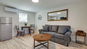 A modern living room with a grey sofa, a wooden coffee table, a small dining table with chairs, a refrigerator, and a wall-mounted mirror. - 3 Mi to Beach & Pier: Pet-Friendly Cape Cod Apt! (Wareham)
