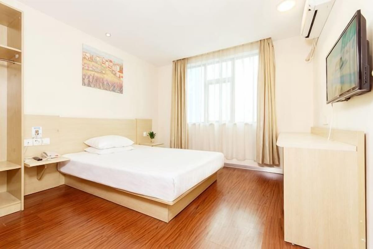 A hotel room with a bed, a desk, a chair, a wardrobe, a window with curtains, and a painting on the wall.