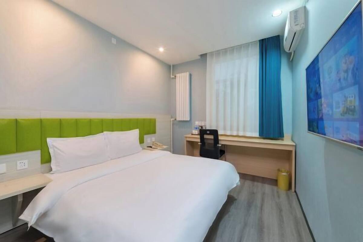Quarto de hotel com cama, escrivaninha, cadeira, janela com cortinas e ar condicionado.