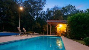 A rectangular swimming pool with a tiled edge, surrounded by a concrete deck. There are lounge chairs along the poolside, and a covered area with a lamp post in the background. - Casa Fofo Family House (San Carlos)