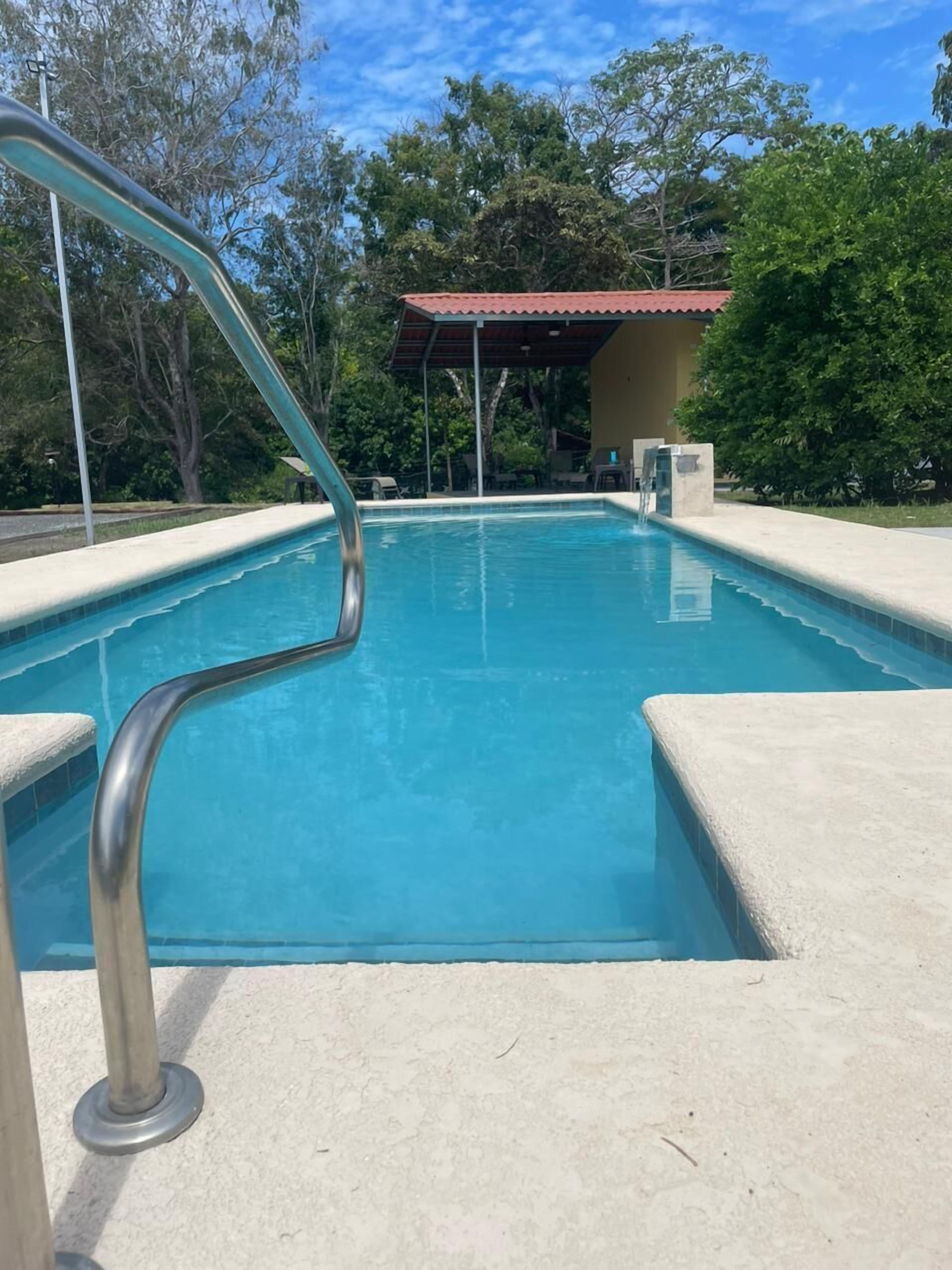 Una piscina con barandilla de metal, rodeada de una solana de hormigón y árboles.