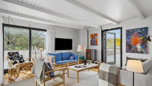 A spacious living room with a blue sofa, a wooden rocking chair, a grey sofa, and a coffee table. There is a TV mounted on the wall, a fireplace, and large windows offering a view of the outdoors. - Pet friendly home in Saltum with WiFi (Saltum)