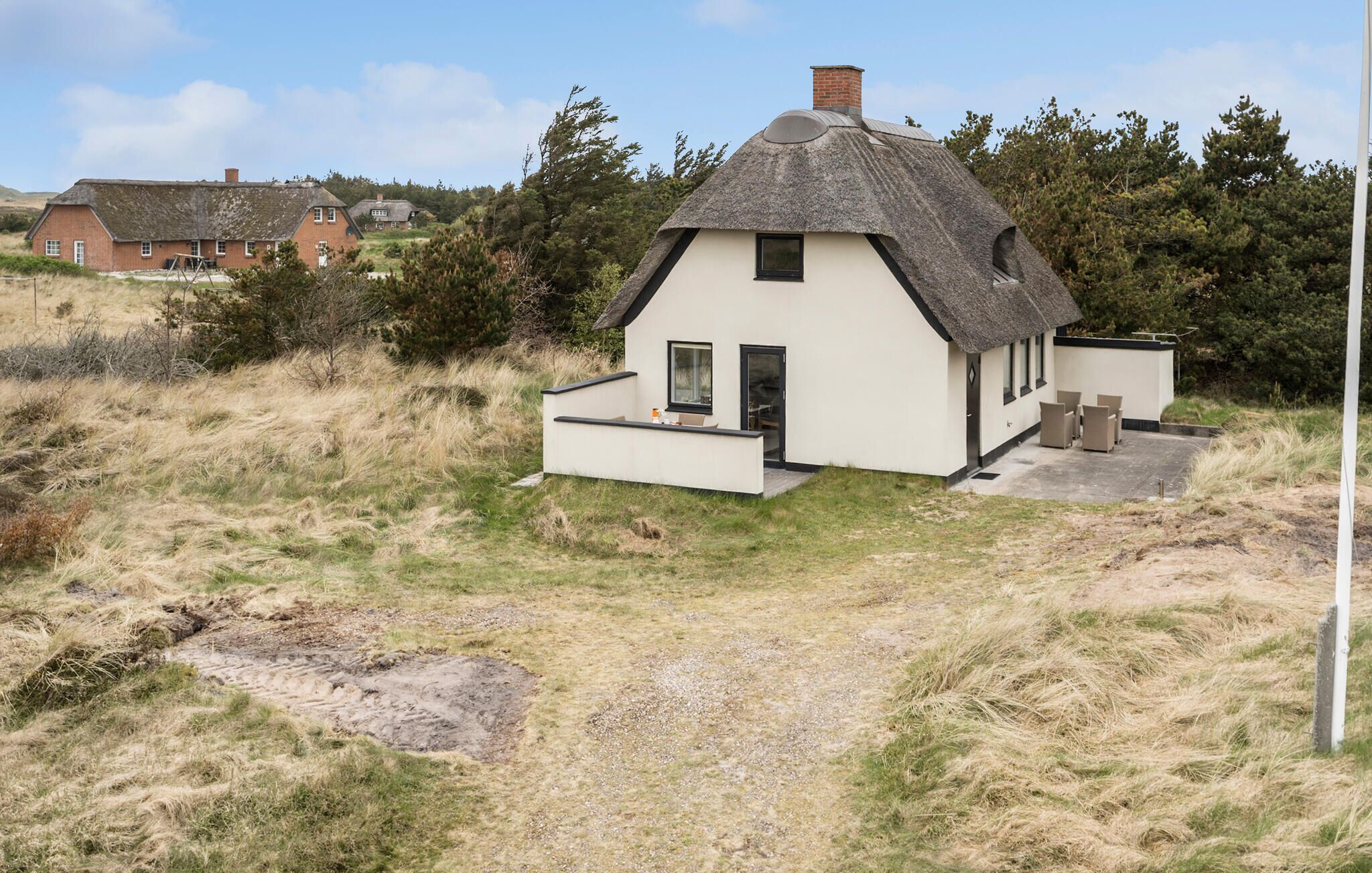 Une maison blanche avec un toit de chaume, un petit porche avec des chaises et une zone pavée devant.