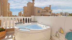 Eine Dachterrasse mit Whirlpool, weißer Brüstung und Blick auf Gebäude im Hintergrund.