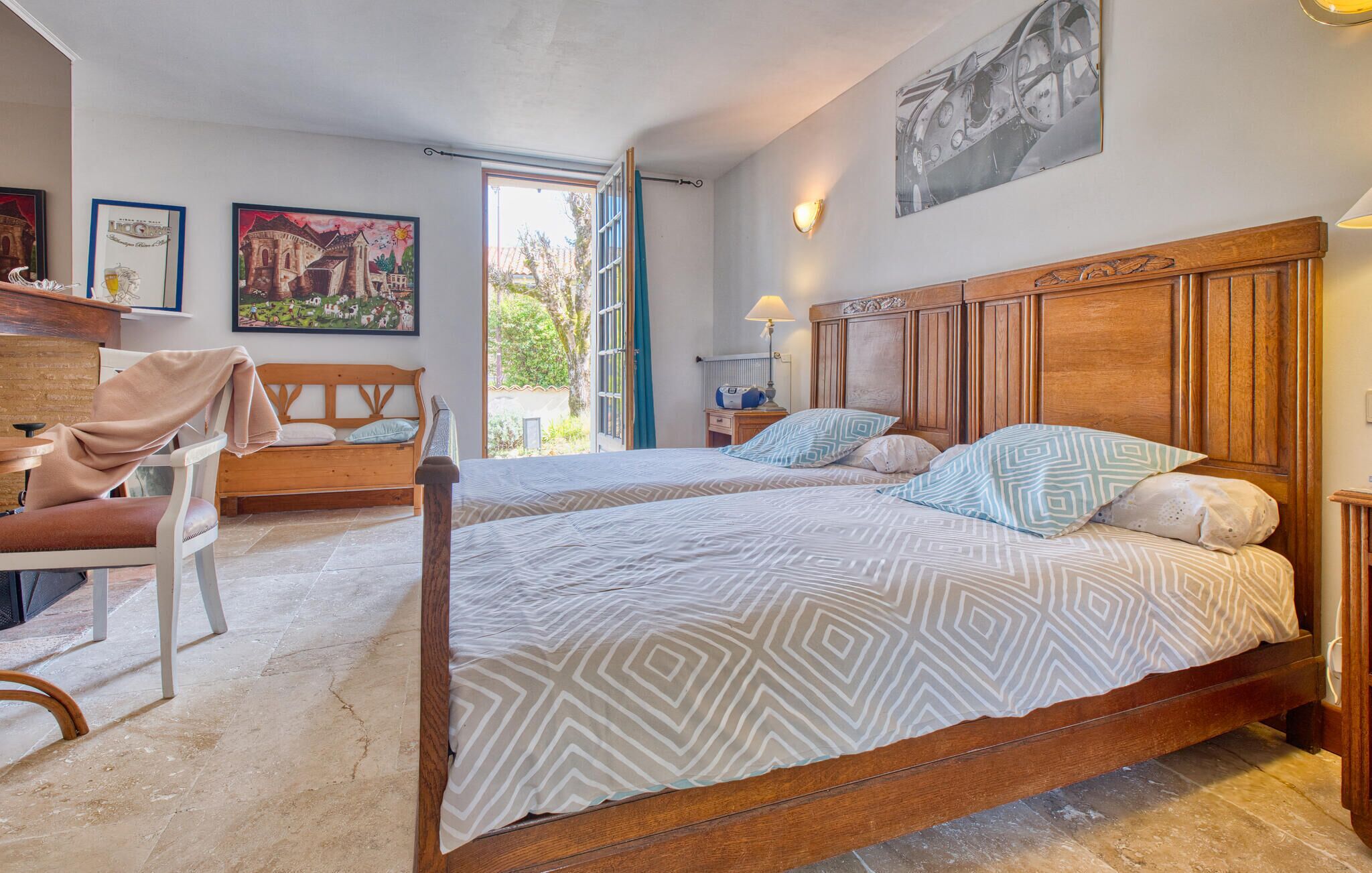 Een slaapkamer met twee bedden, een houten hoofdbord, een raam met uitzicht op groen en een tafel met stoel.