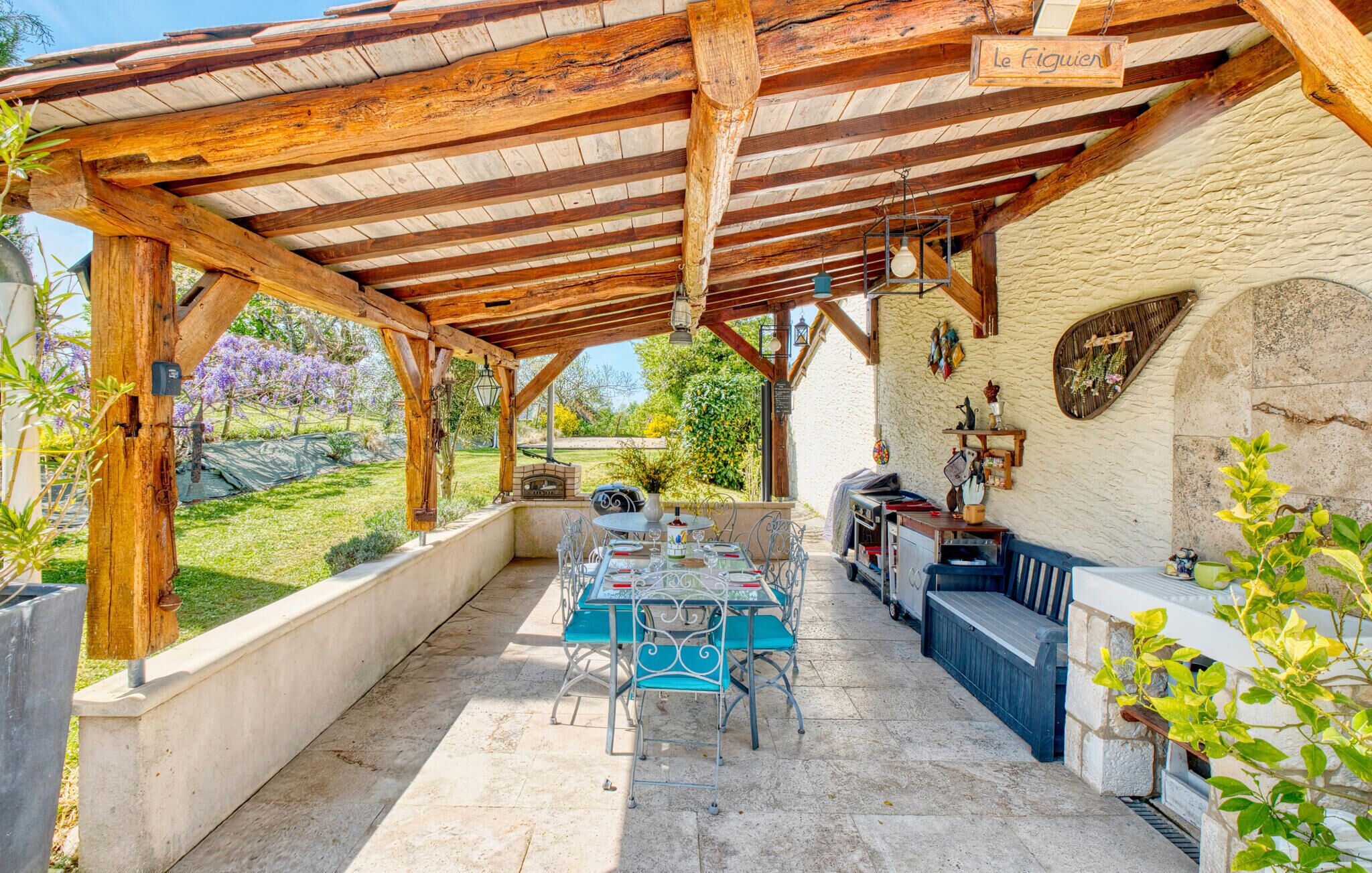 Un patio exterior con una pérgola de madera, una mesa con sillas, una parrilla de barbacoa y un muro de piedra con un letrero que dice "Le Figuiere".