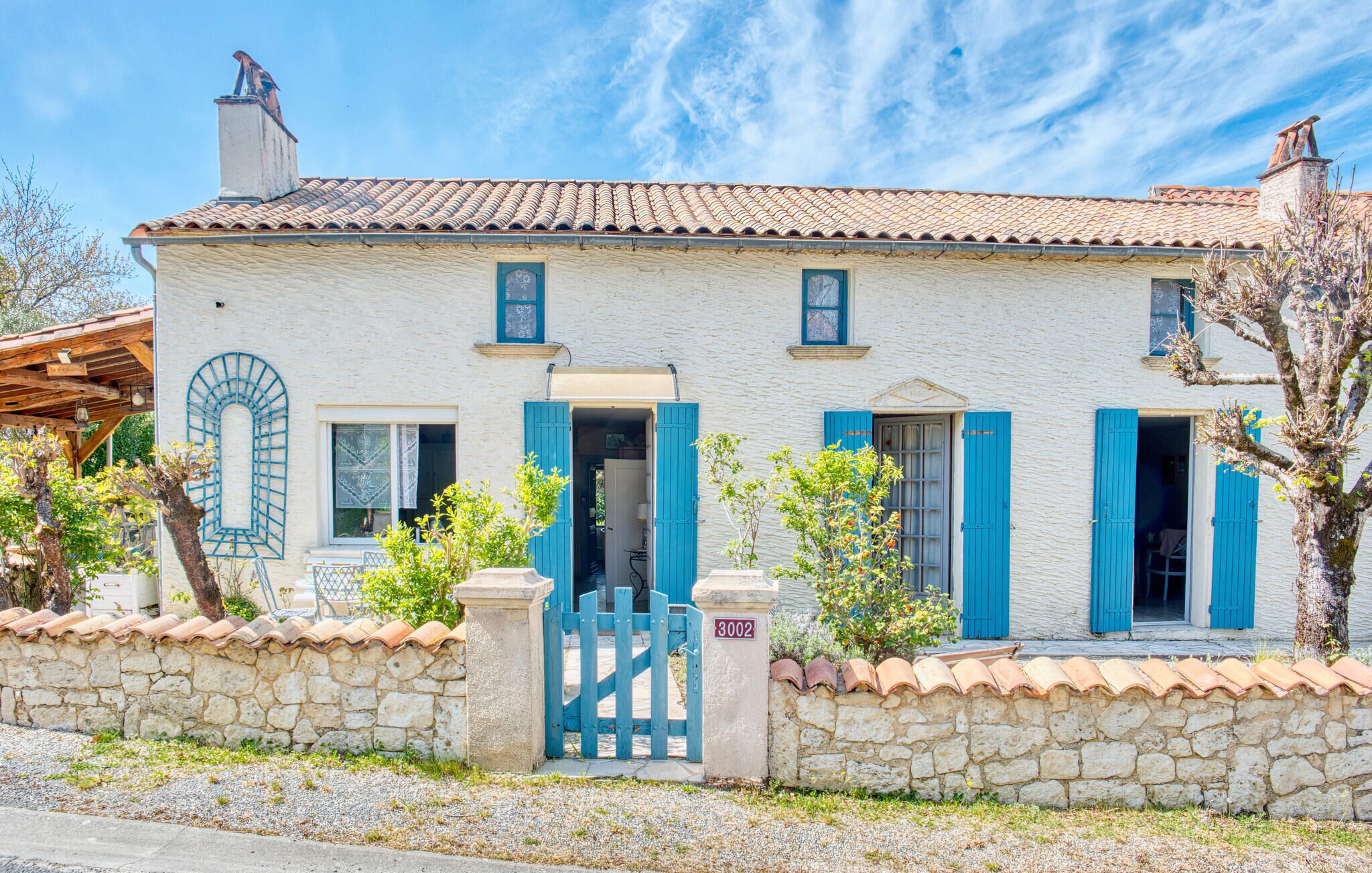 A two-story stone building with a tiled roof, blue shutters, and a stone wall with a gate.