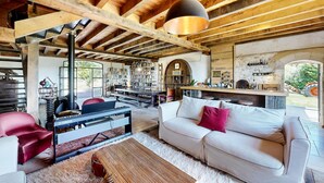 A spacious living area with a high ceiling, exposed wooden beams, a large sofa, a red chair, a Yamaha piano, a bookshelf, and a kitchen area visible through an open archway.