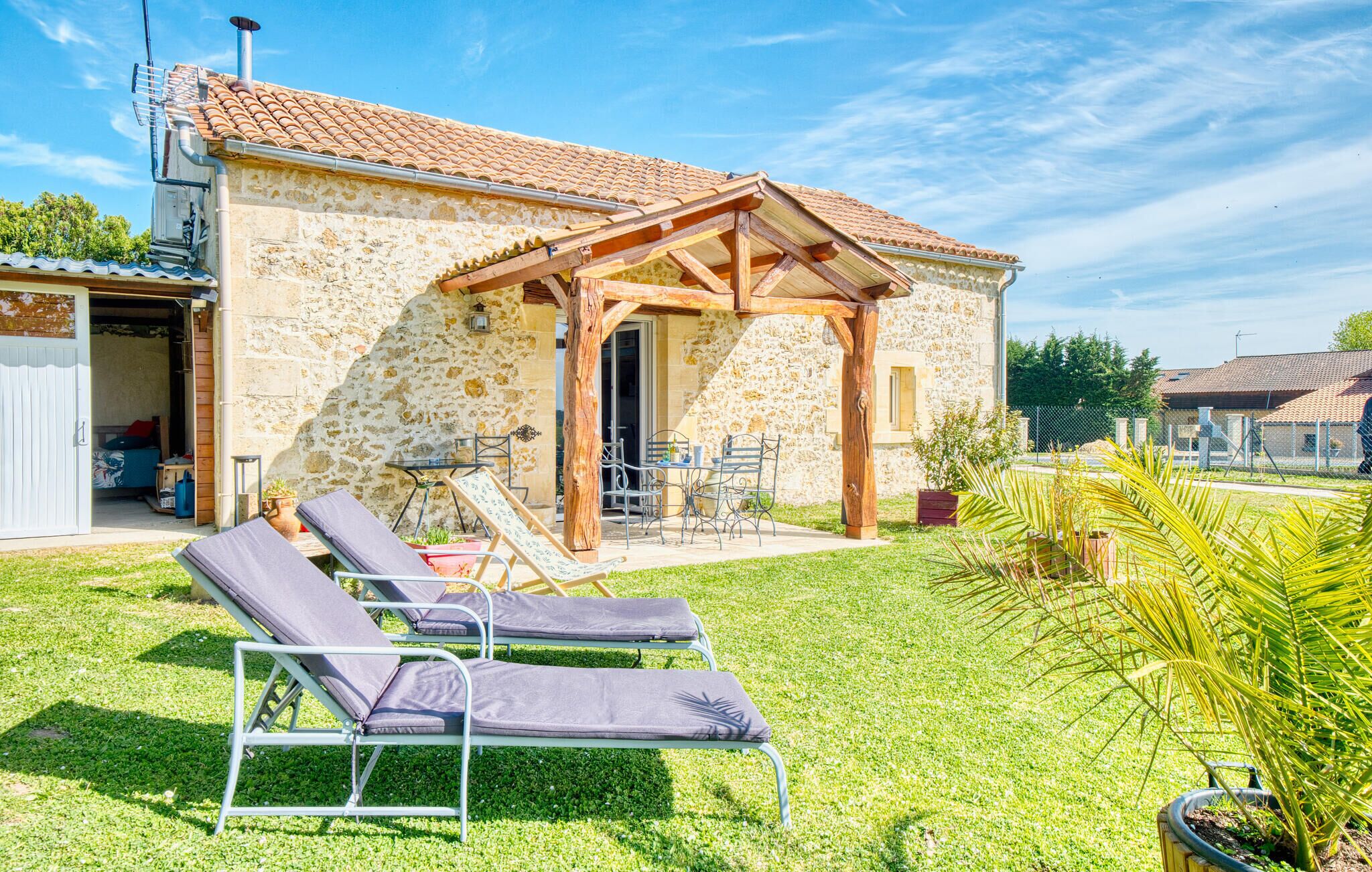 A stone house with a tiled roof, a wooden pergola, and a patio with lounge chairs.