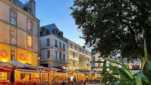 Une rue pavée bordée de restaurants avec des tables en terrasse, entourée de bâtiments historiques dont les panneaux indiquent la présence d’un hôtel et d’un château.