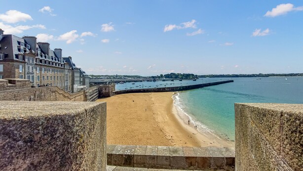 Une vue sur la côte, avec une plage de sable, un mur de pierre et une rangée d’immeubles à étages et dotés de balcons.
