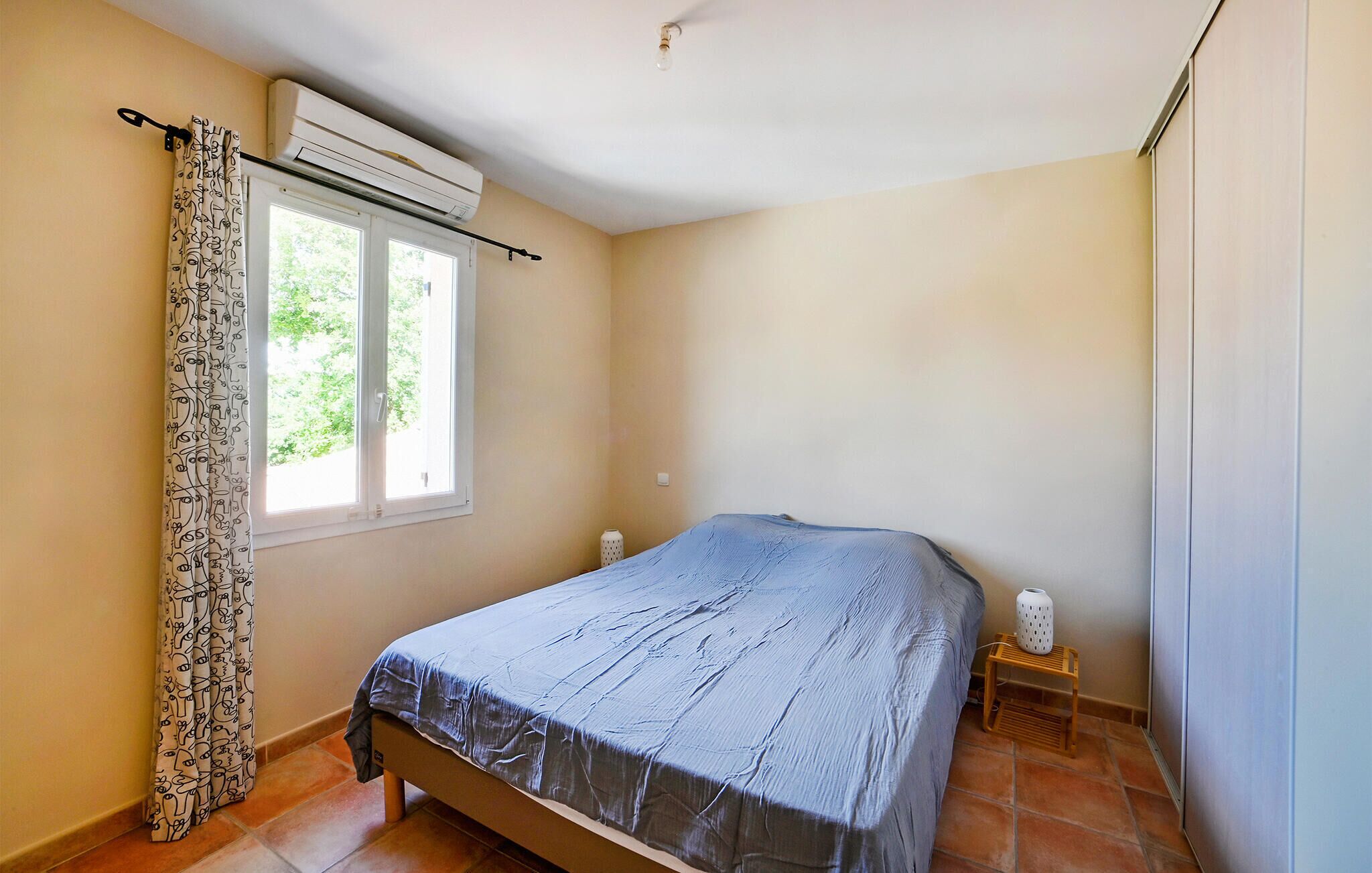 Een slaapkamer met een bed, een airconditioner, een raam met gordijnen en een nachtkastje met een lamp.