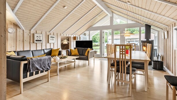 Un vaste espace de vie doté d’un haut plafond, de poutres apparentes, d’un ensemble de canapés, d’une table à manger avec des chaises, d’une cheminée et de grandes fenêtres offrant une vue sur l’extérieur.