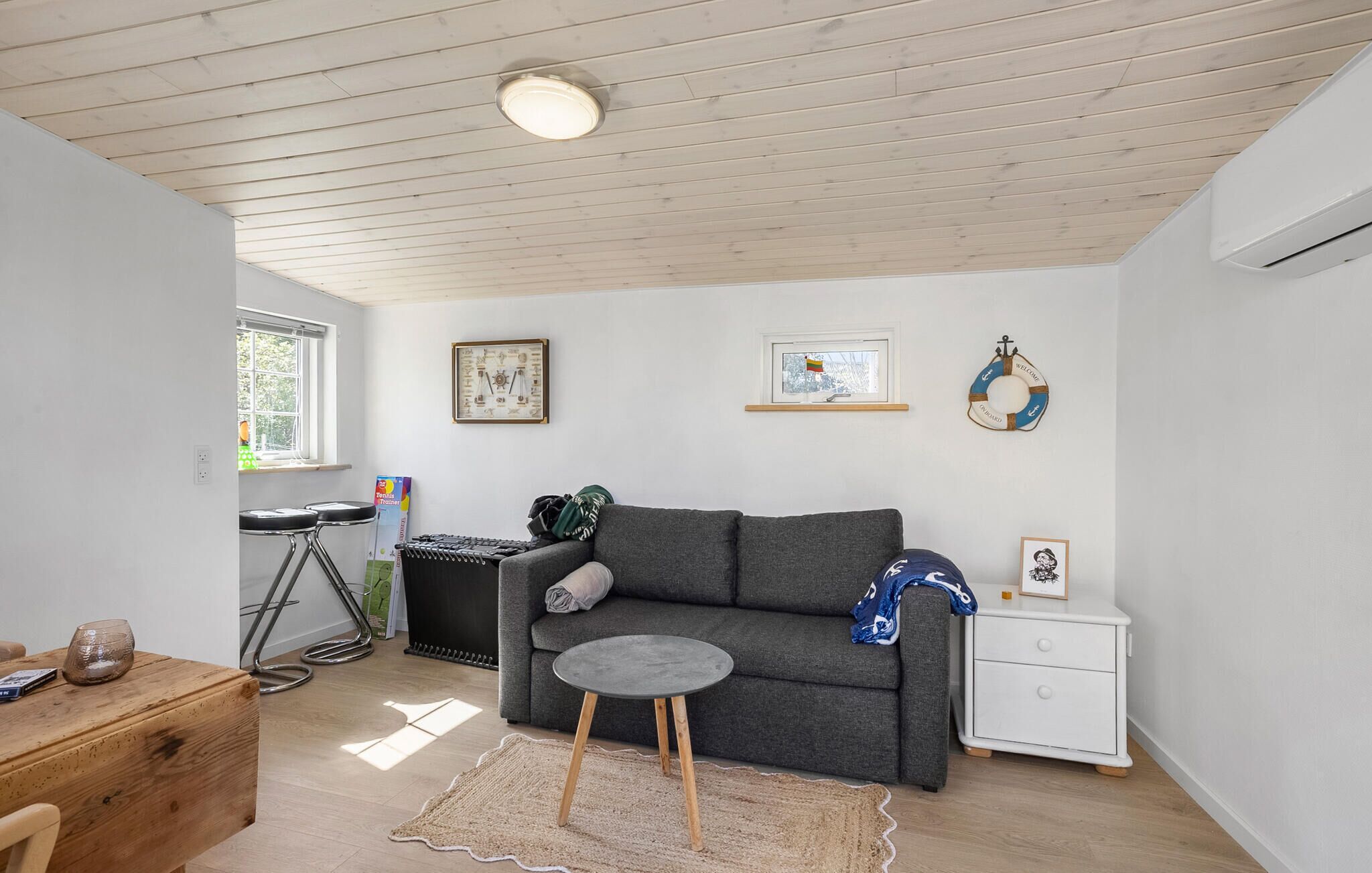 A cozy living room with a grey sofa, a small round table, a white nightstand, and a wooden desk with a tennis racket.