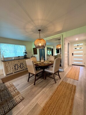 A dining area with a round table, chairs, a hanging light fixture, a cabinet with drawers, and a window with blinds.