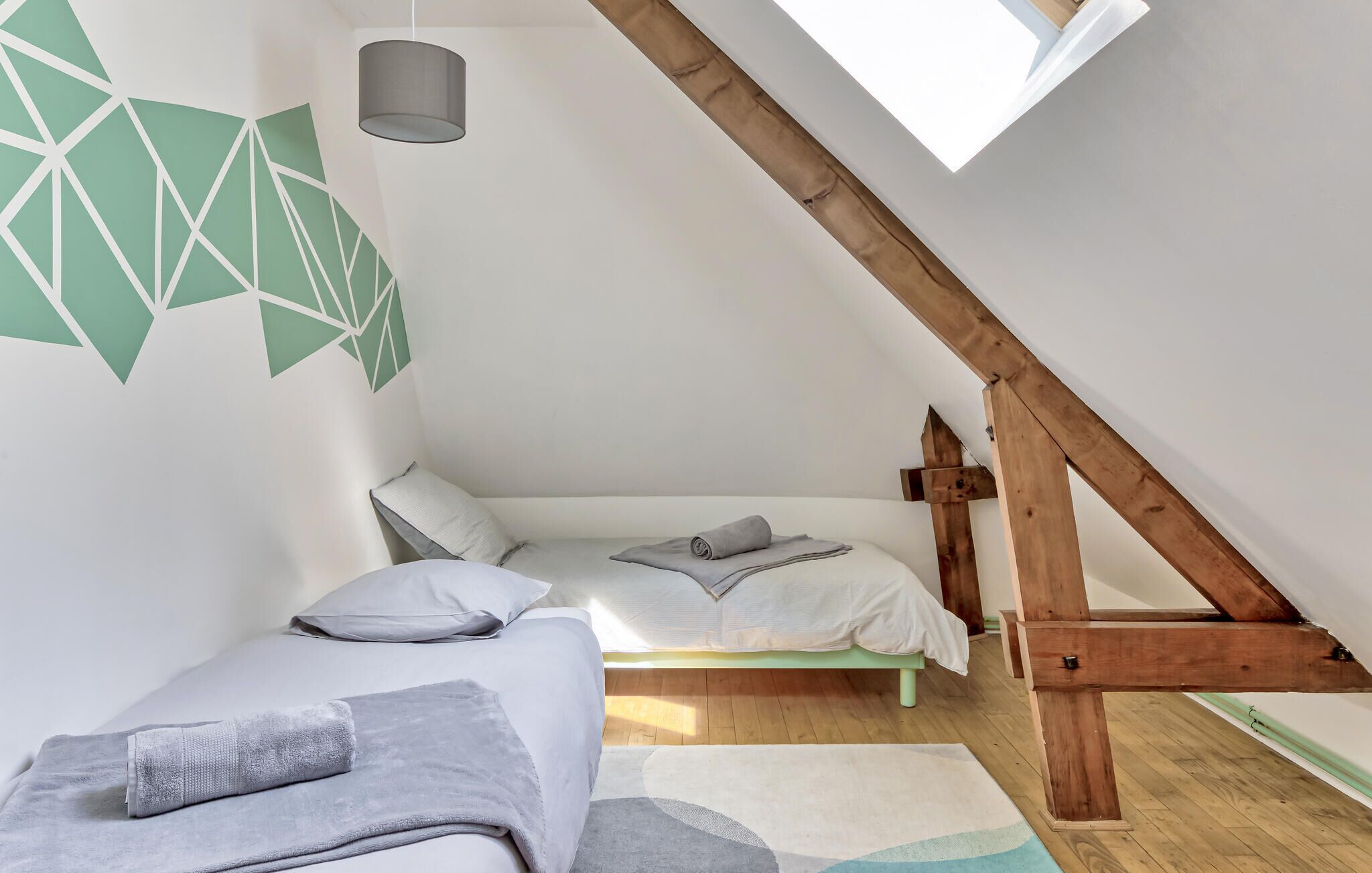Une chambre mansardée chaleureuse, avec un plafond en pente, une structure en poutres apparentes, une lucarne et un motif géométrique sur le mur.