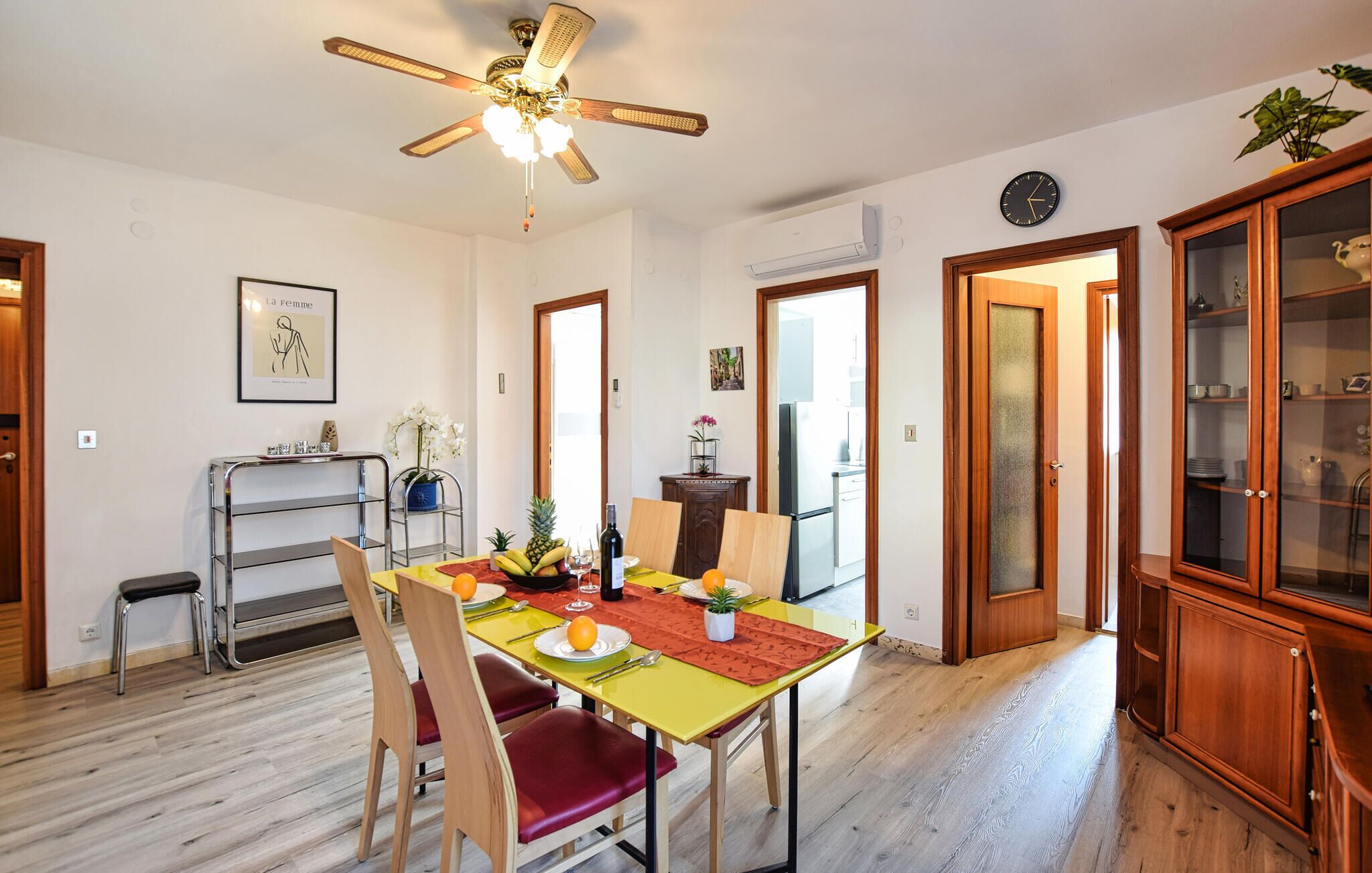 Un comedor con mesa de madera, sillas y ventilador de techo. Se ve una cocina a través de una puerta abierta.