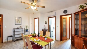 Un coin repas avec une table en bois, des chaises et un ventilateur de plafond. On peut apercevoir la cuisine à travers une porte ouverte.