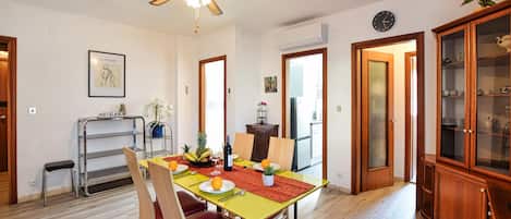 Uma sala de jantar com mesa de madeira, cadeiras e ventilador de teto. Cozinha visível por uma porta aberta.