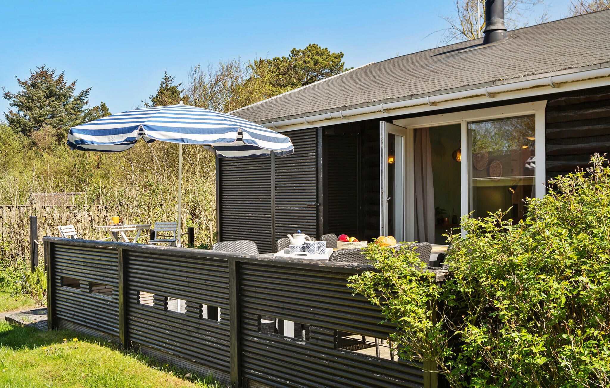 En træterrasse med en stribet parasol, et bord med stole og et vindue med gardiner.