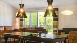 A dining area with a wooden table and chairs, two pendant lights, and a view of a green outdoor space through large windows. - Awesome home in Oksbøl with sauna (Oksbøl)