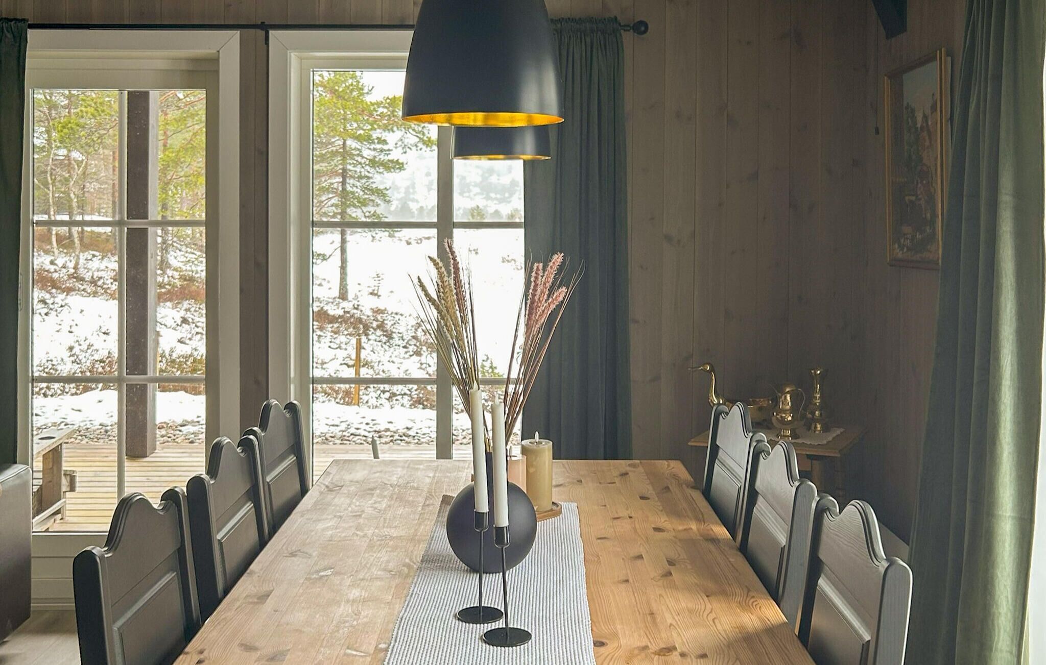 A dining room with a wooden table, chairs, a black pendant light, and a view of a snowy landscape through large windows.