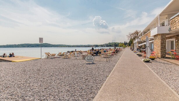 A pebble beach with lounge chairs and tables, a pathway, and a building with outdoor seating. - Pet friendly apartment (Soiano del Lago)