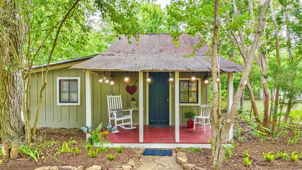 A rustic house with a porch, two white chairs, a red door, and a heart-shaped decoration.