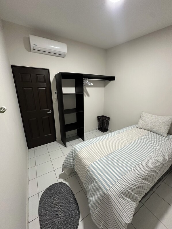 A bedroom with a bed, a black wardrobe, a hanging shelf, a trash bin, and an air conditioning unit.