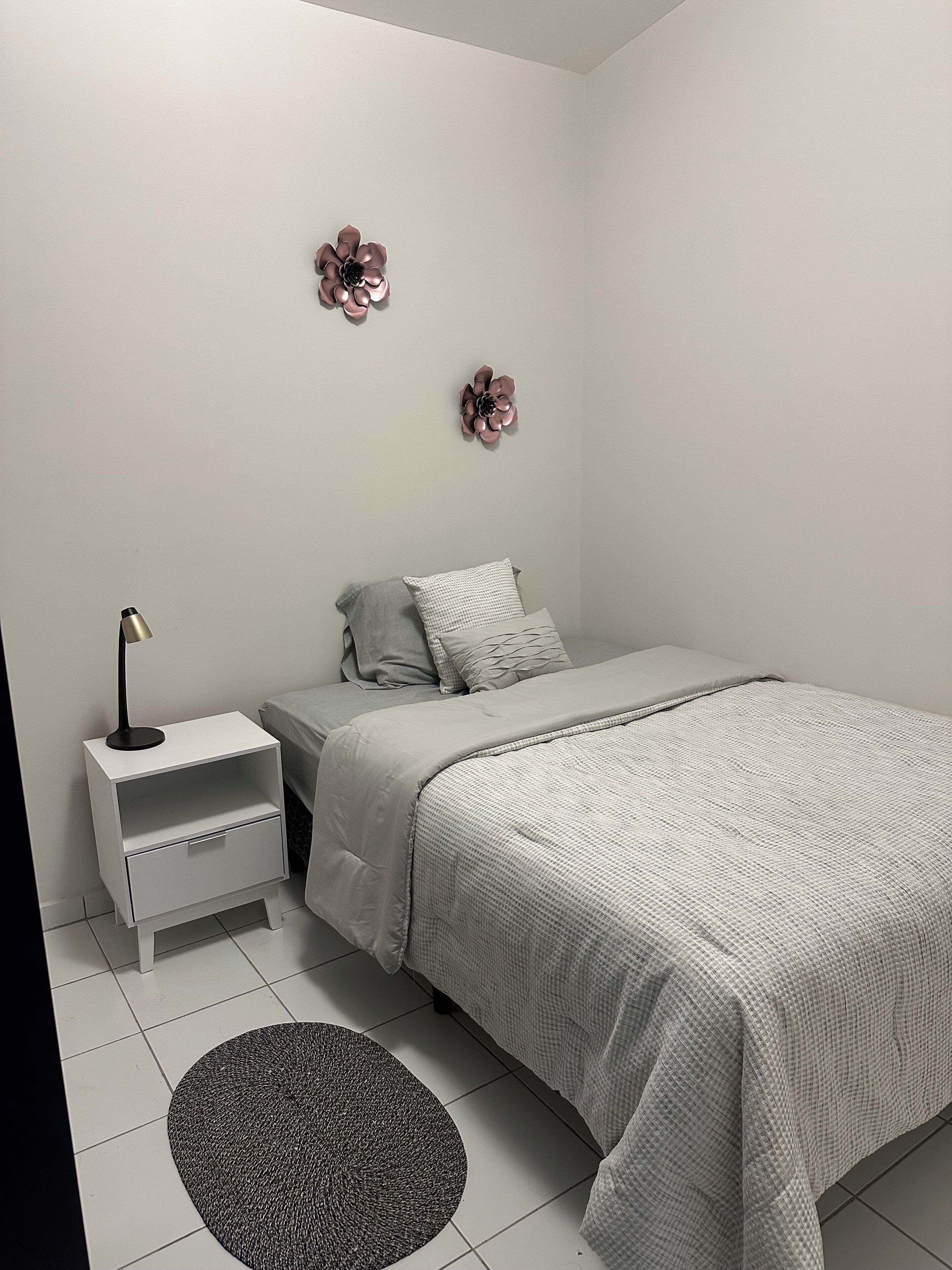 A bedroom with a bed, nightstand, lamp, decorative flowers, and a rug.