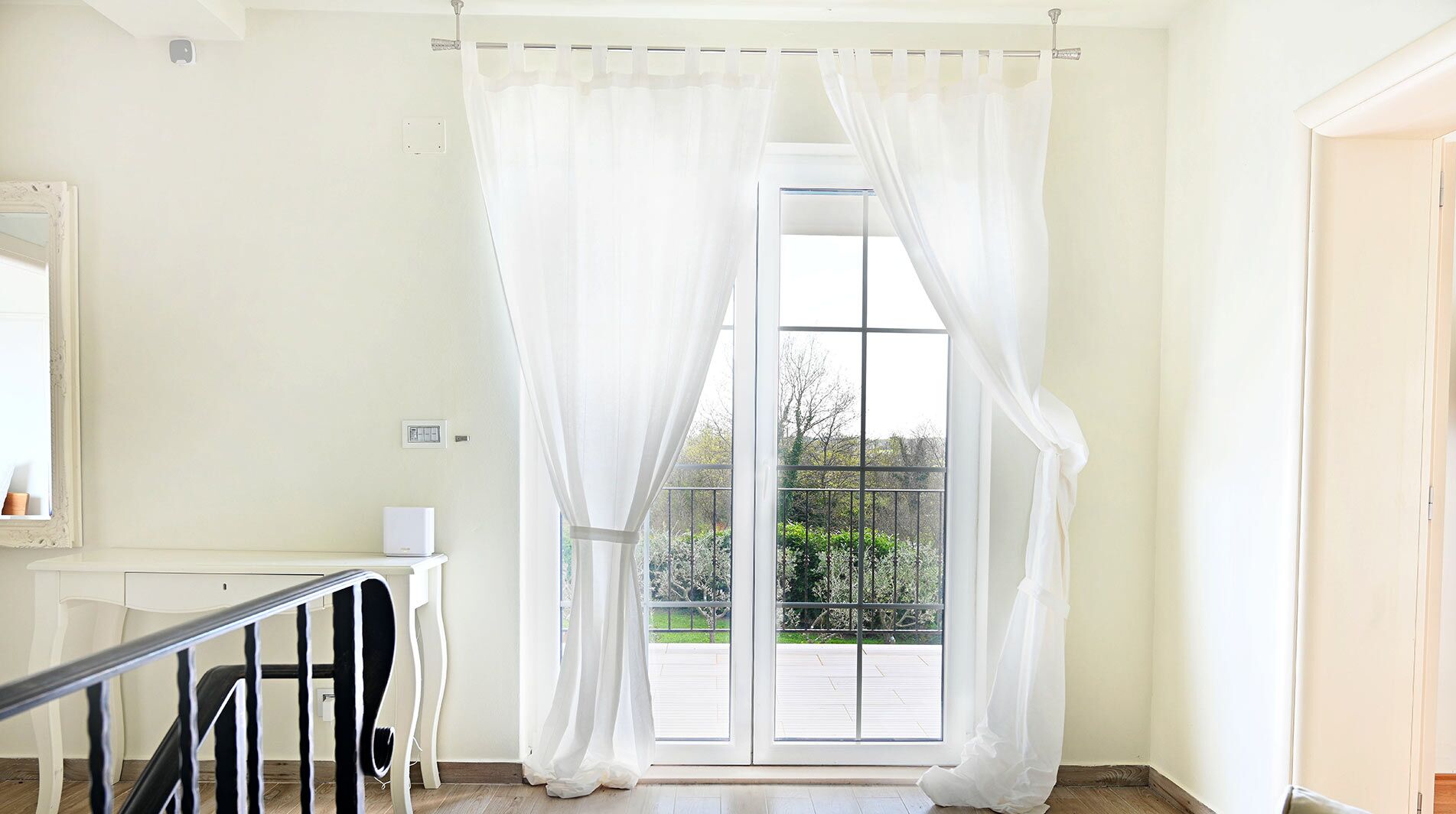 A room with a white door and curtains, a small table with a chair, and a view of a balcony with greenery.