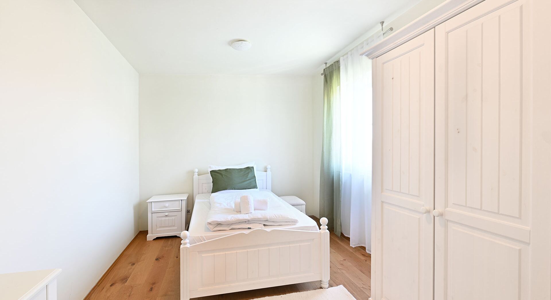 A bedroom with a single bed, a nightstand, a wardrobe, and a window with curtains.