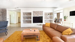 Een moderne woonkamer met een stenen muur, een flatscreen televisie, een houten salontafel en een bruine bank met gele kussens.