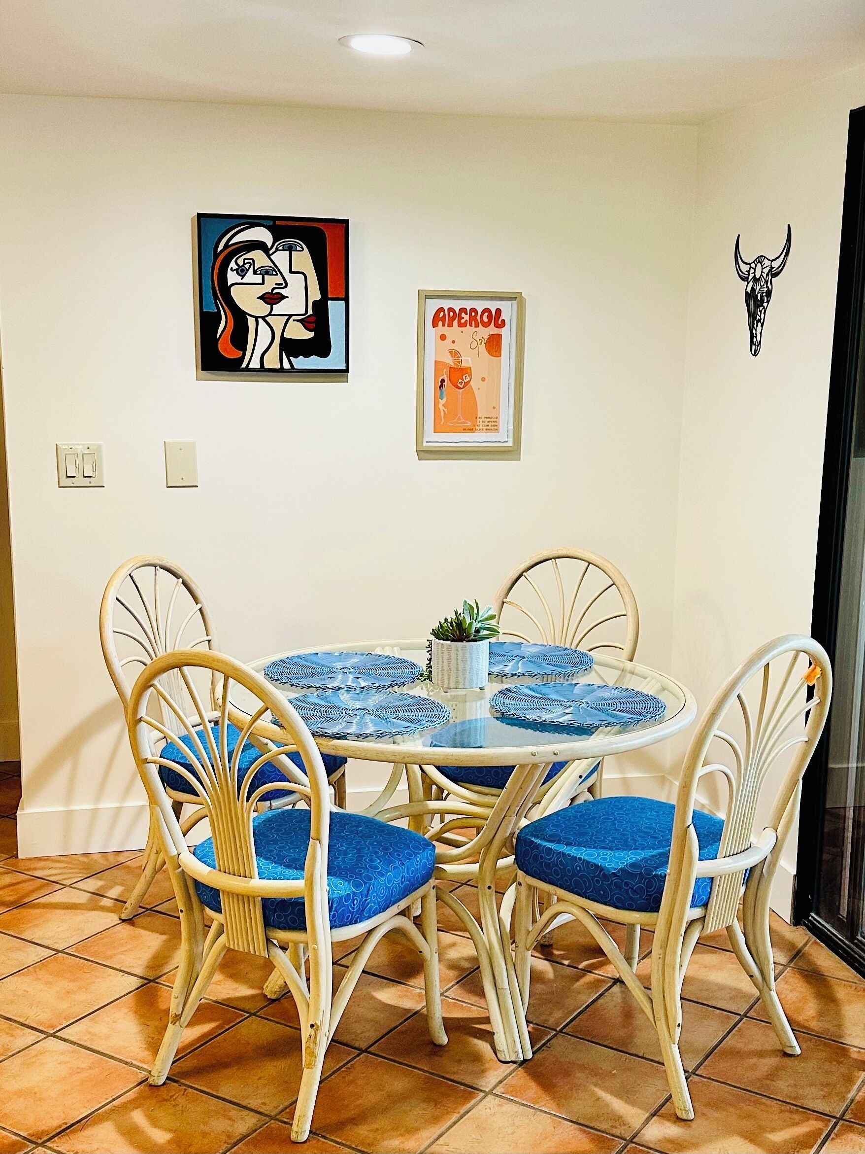 Een eethoek met een ronde tafel en vier stoelen, elk voorzien van blauwe kussens. Twee decoraties aan de muur: een is een ingelijste poster met tekst en een illustratie, de ander is een zwart-wit afbeelding van een koeienhoofd.
