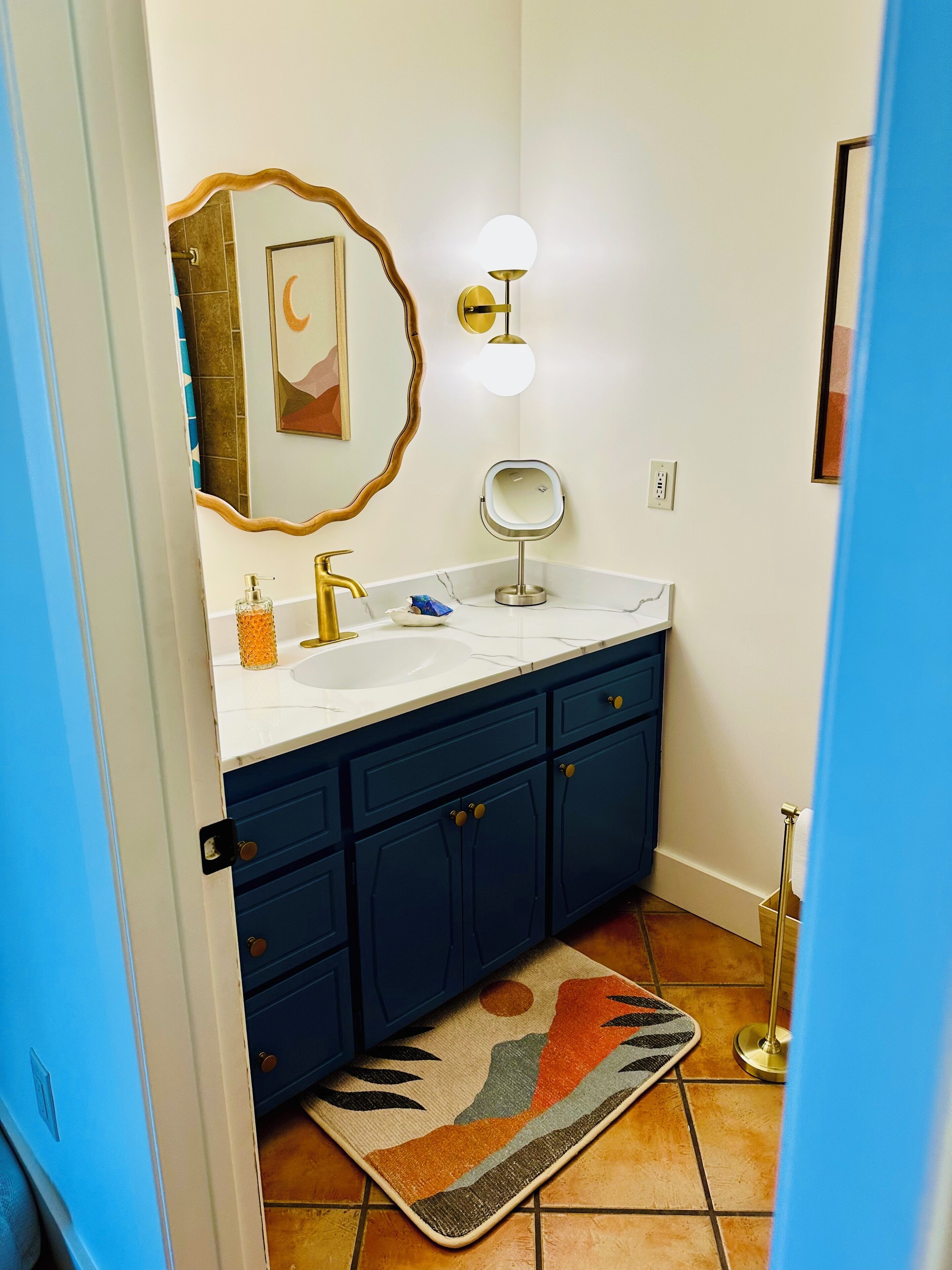 A bathroom with a white sink, blue cabinets, a decorative rug, a wall-mounted mirror, and a light fixture.