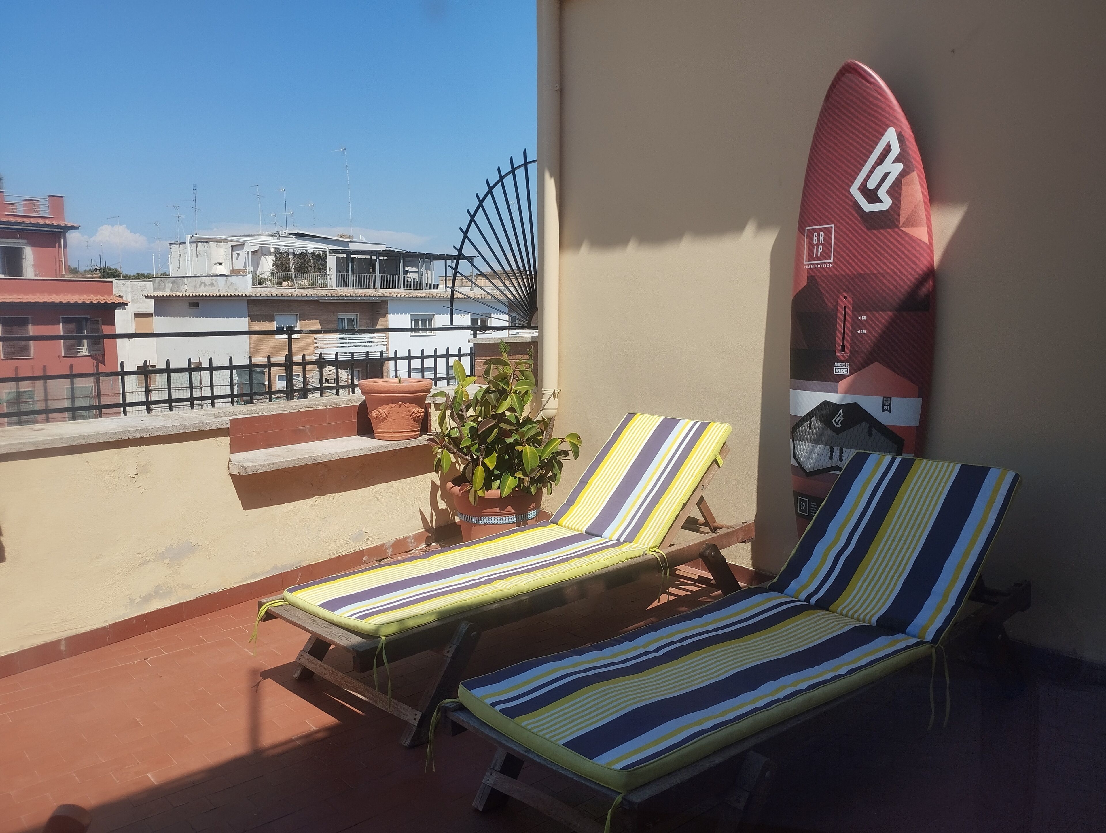 A balcony with two striped lounge chairs, a potted plant, and a surfboard against the wall.