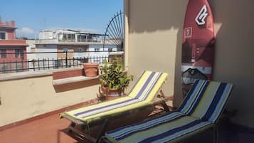 A balcony with two striped lounge chairs, a potted plant, and a surfboard against the wall.