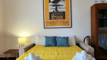 Ein Hotelzimmer mit gelber Bettdecke, zwei Kissen, einem gerahmten Poster an der Wand, einem Holzschrank und einem Nachttisch mit Lampe.