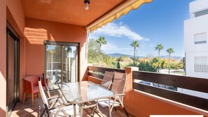 Un balcon avec une table et des chaises, un luminaire suspendu, et une vue sur des palmiers et des montagnes.