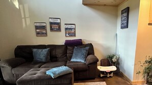 A cozy living room with a sectional sofa, a small ottoman, and a patterned rug. There are wall decorations and a ceiling with exposed beams. - Charming lake cottage overlooking Grand lake (Grove)