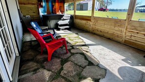 A covered outdoor seating area with stone flooring, wooden walls, and a view of a grassy field. - Charming lake cottage overlooking Grand lake (Grove)