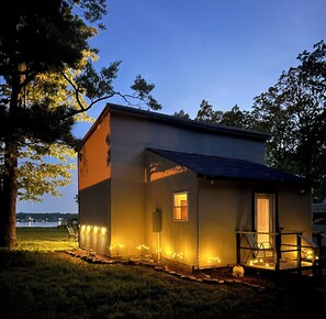 A small, single-story building with a sloped roof, a porch with a railing, and a small window. - Charming lake cottage overlooking Grand lake (Grove)