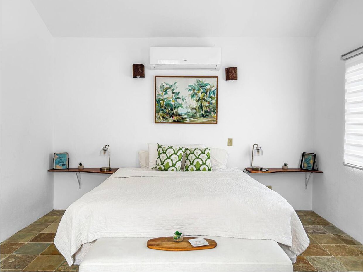 A bedroom with a bed, a nightstand, a wall-mounted air conditioner, a framed painting, and a window with blinds.