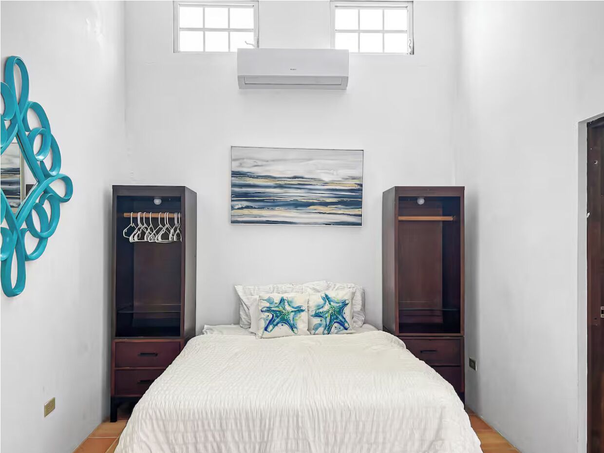 Een slaapkamer met een bed, twee kasten, een wandairconditioner, een raam en een schilderij.