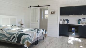 Une chambre à coucher comprenant un lit, une table de chevet, une lampe et une porte donnant sur la cuisine.